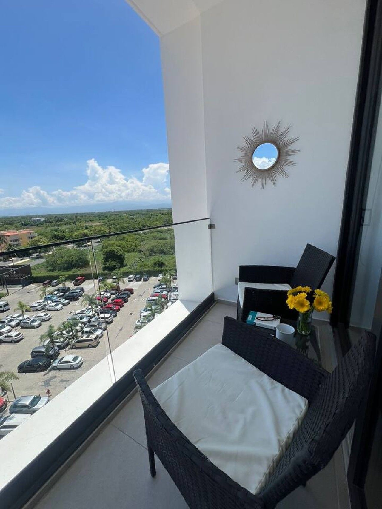 Un balcon donnant sur un parking et sur un espace verdoyant, un miroir en forme de soleil et une chaise en osier avec un coussin.