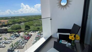 Un balcon donnant sur un parking et sur un espace verdoyant, un miroir en forme de soleil et une chaise en osier avec un coussin.