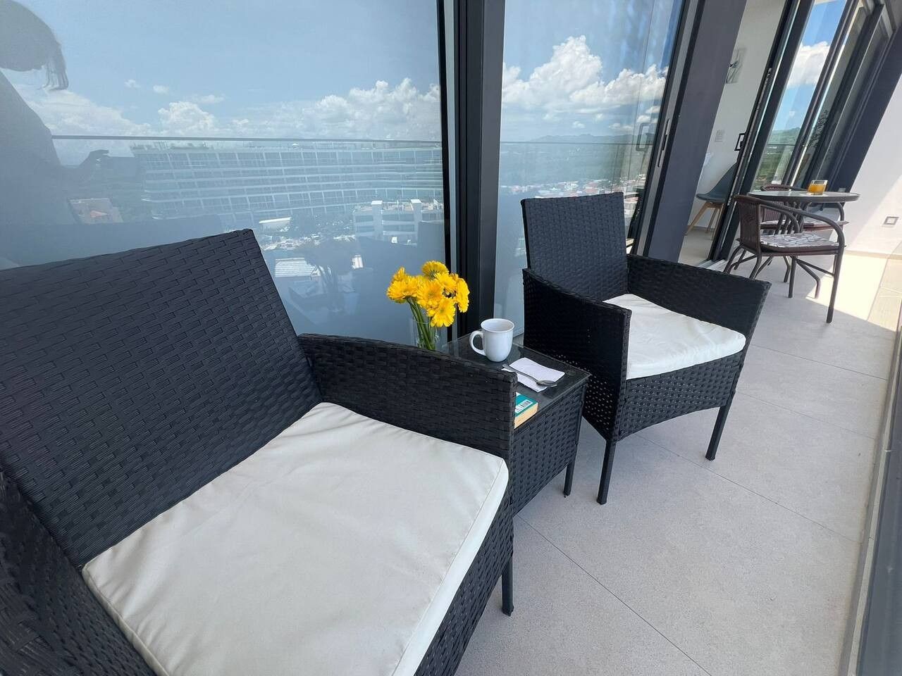 A balcony with wicker furniture, a vase with yellow flowers, and a clear view of the cityscape.