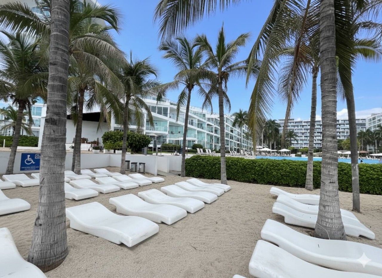 Zona de playa con sillones blancos dispuestos en filas, palmeras y un edificio con piscina al fondo.