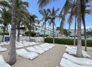 Zona de playa con sillones blancos dispuestos en filas, palmeras y un edificio con piscina al fondo.