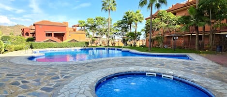 A swimming pool with stone tiles, surrounded by palm trees and buildings.