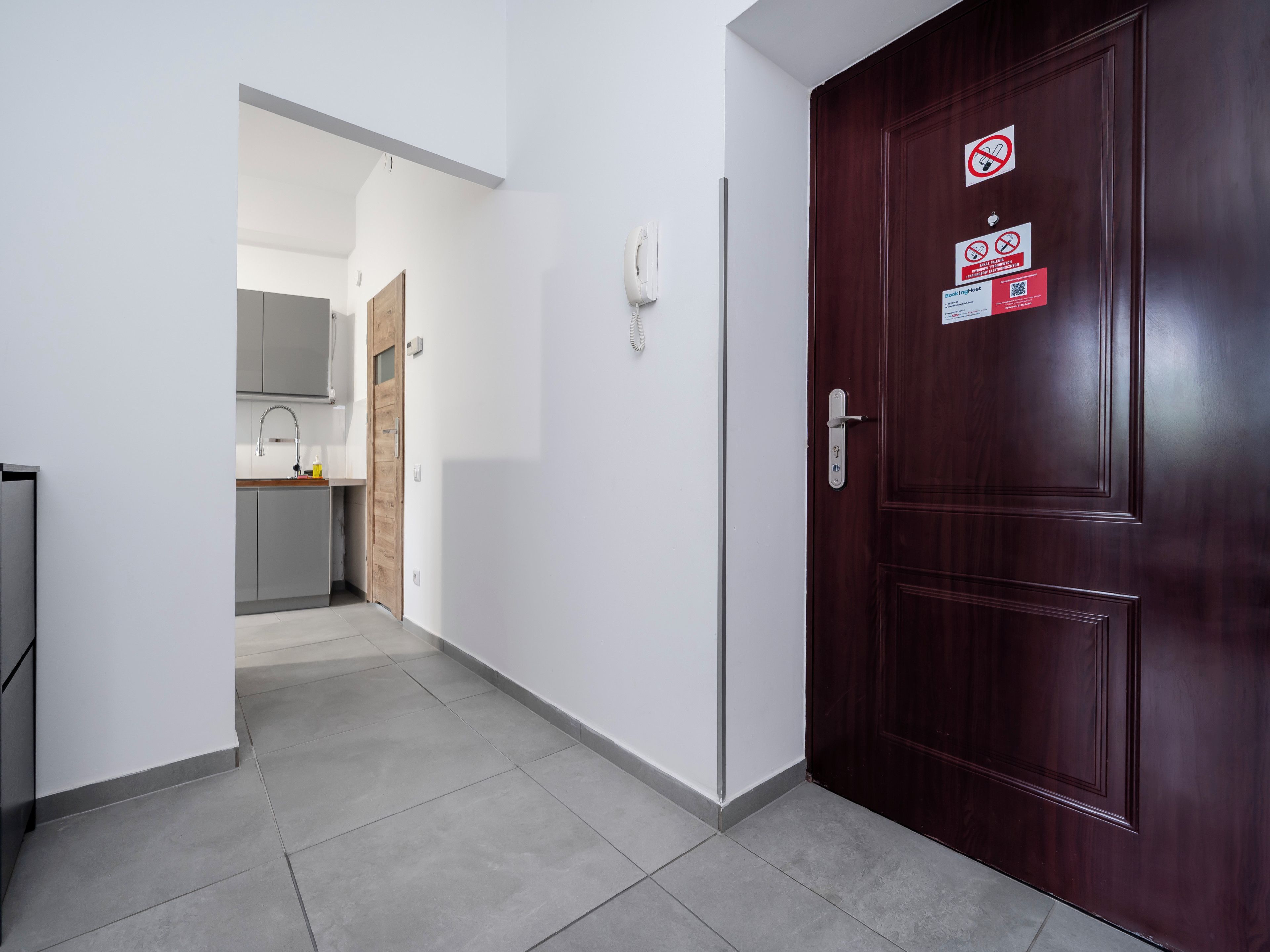 A hallway with a wooden door, a kitchen area, and a white wall with a phone and signs.
