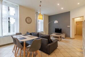 A modern living room with a sofa, dining table, chairs, a TV, and a clock on the wall. - Zwycięstwa Comfort Apartment (Gliwice)