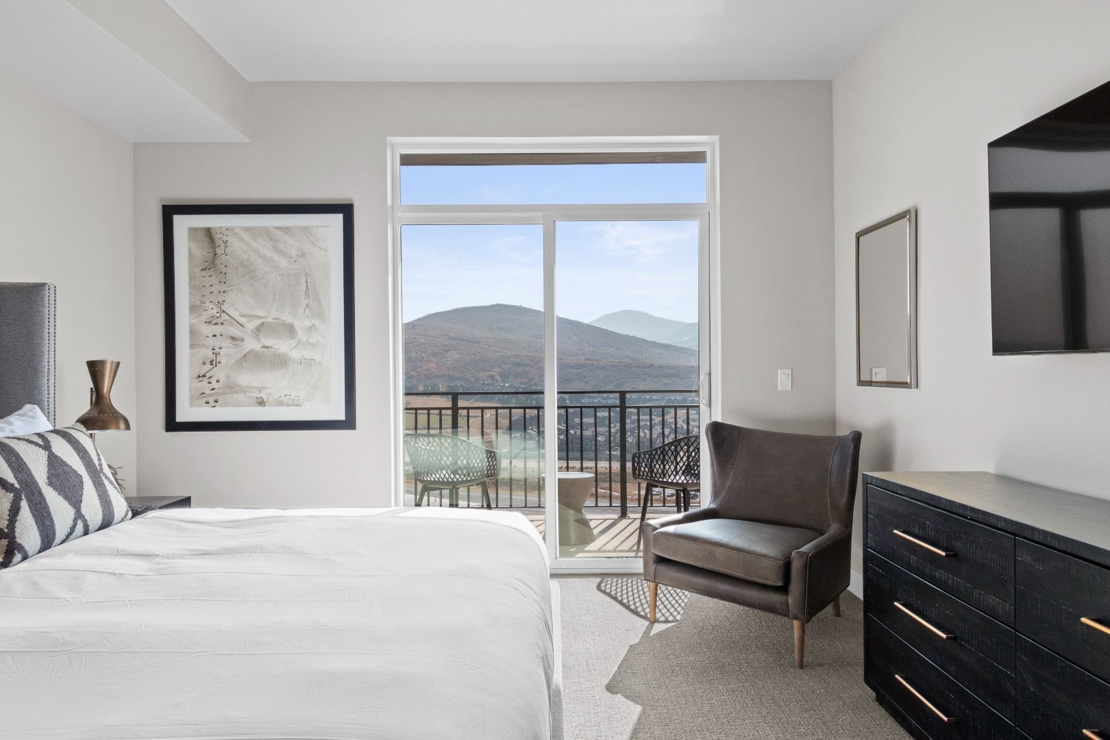 A bedroom with a large bed, a chair, a dresser, and a balcony with a view of mountains.