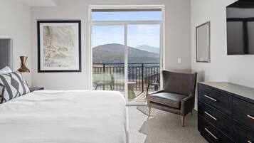 A bedroom with a large bed, a chair, a dresser, and a balcony with a view of mountains.