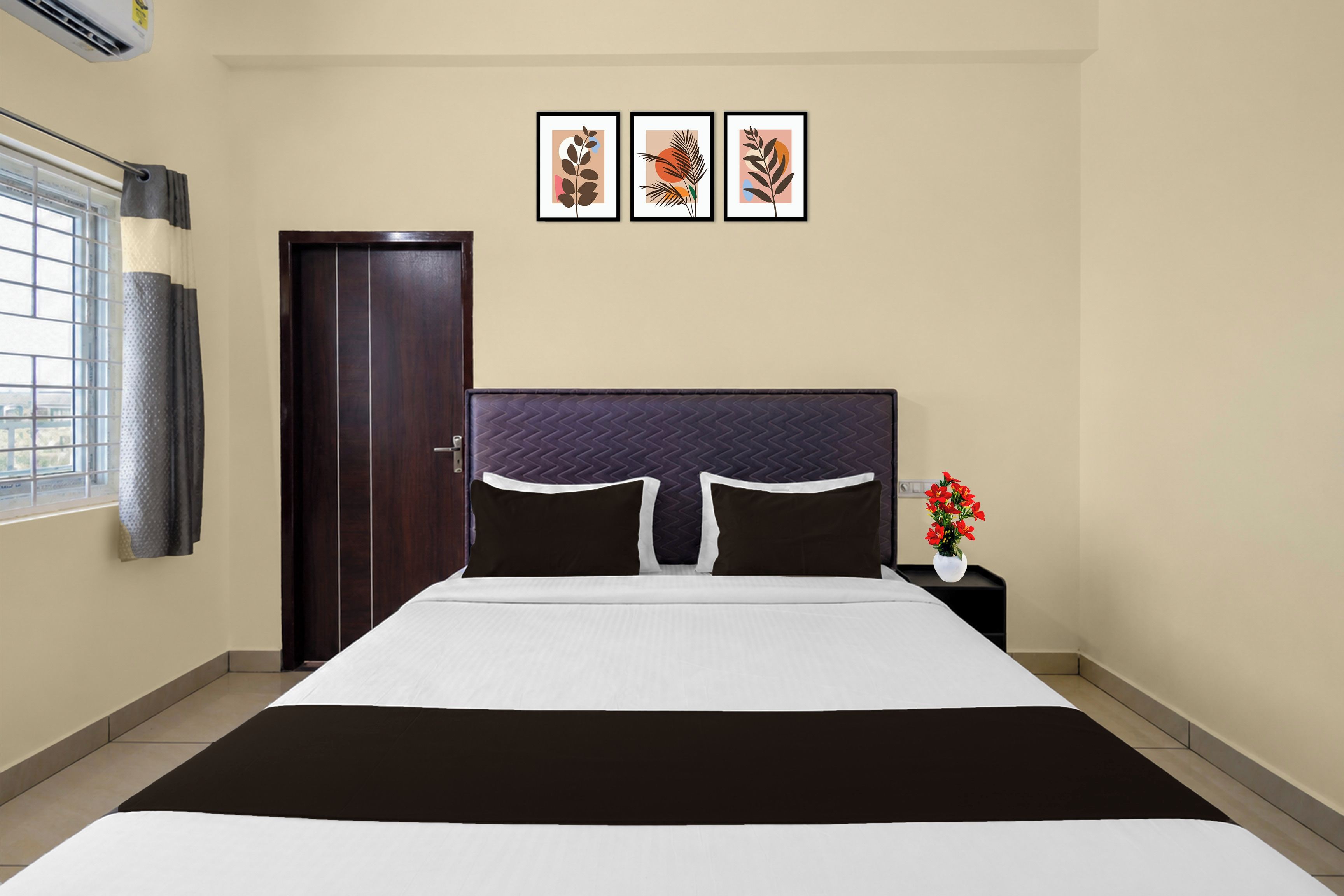 Quarto de hotel com uma cama, mesa de cabeceira, um vaso com flores e três quadros na parede.