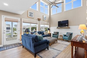 A spacious living room with a large window, a ceiling fan, and a flat-screen TV mounted on the wall. - Waterfront 5br/4.5ba Canal Home Close to The Beach (Ocean Isle Beach)