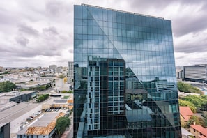 Un edificio moderno de vidrio con fachada en forma de entramado, rodeado de otras construcciones urbanas y un estacionamiento.