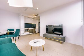 Een moderne woonkamer met een flatscreen televisie, fauteuils bekleed met blauwgroen, een witte salontafel, en een keuken met witte kasten en apparatuur.