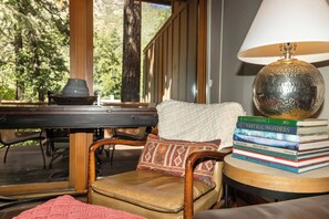A cozy reading nook with a leather chair, a stack of books, a lamp, and a view of a forested area. - Sedona Oak Creek Villa -w/ AC & Heat (Sedona)