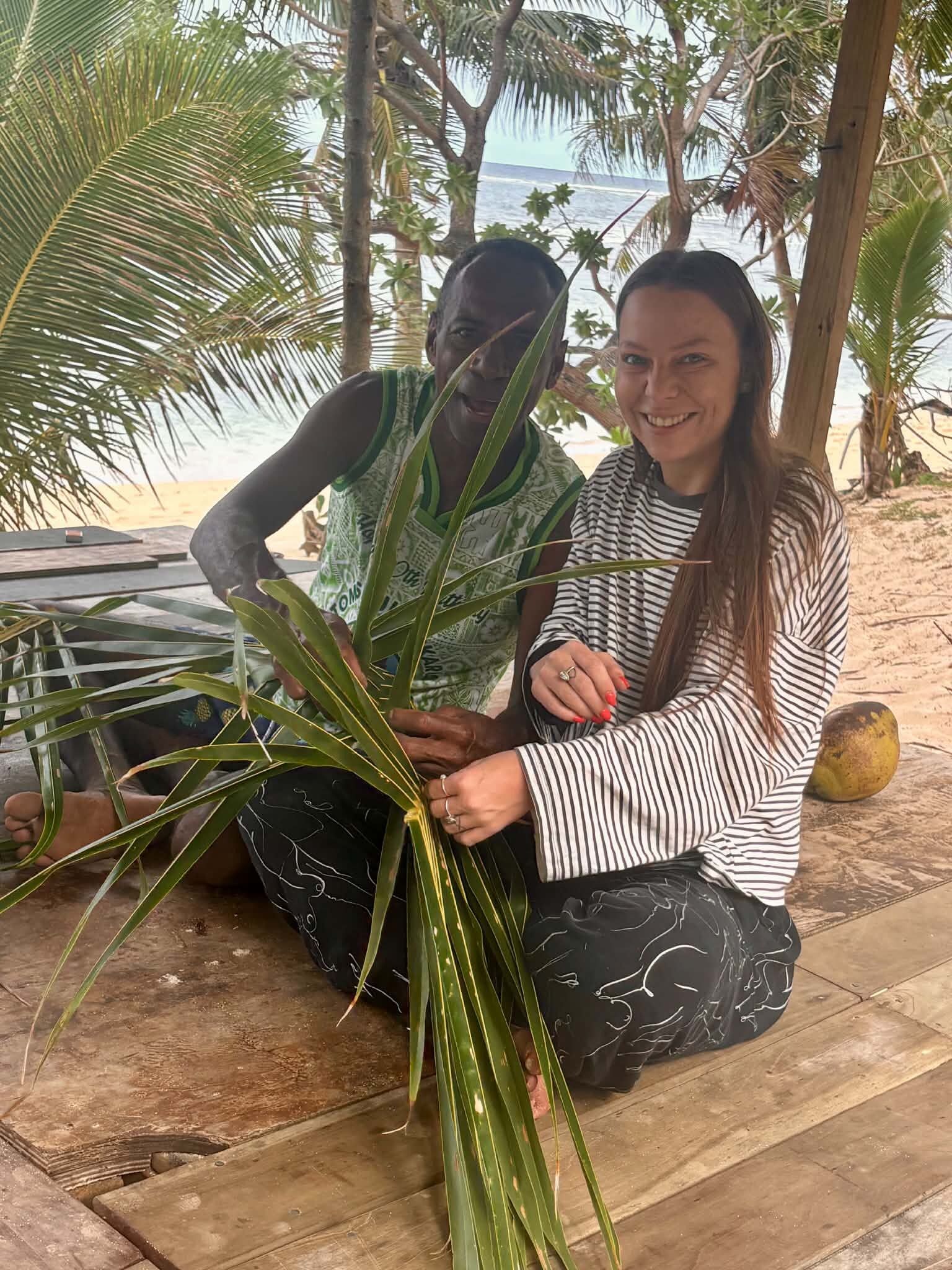 Two people sitting on a wooden deck with palm trees and a beach in the background.