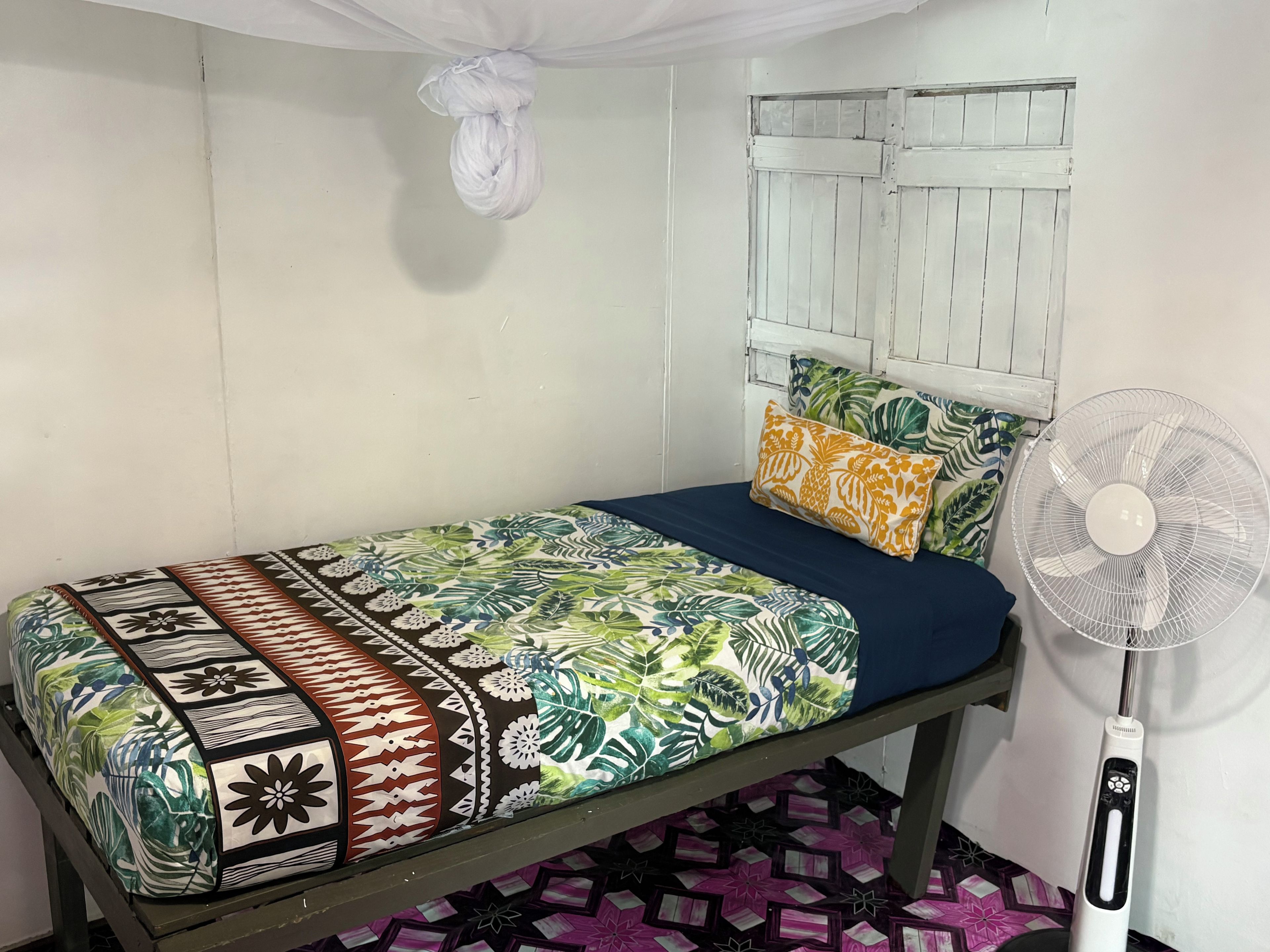 A single bed with a patterned bedspread and pillows, a white fan, and a window with shutters.