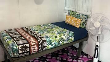 A single bed with a patterned bedspread and pillows, a white fan, and a window with shutters.