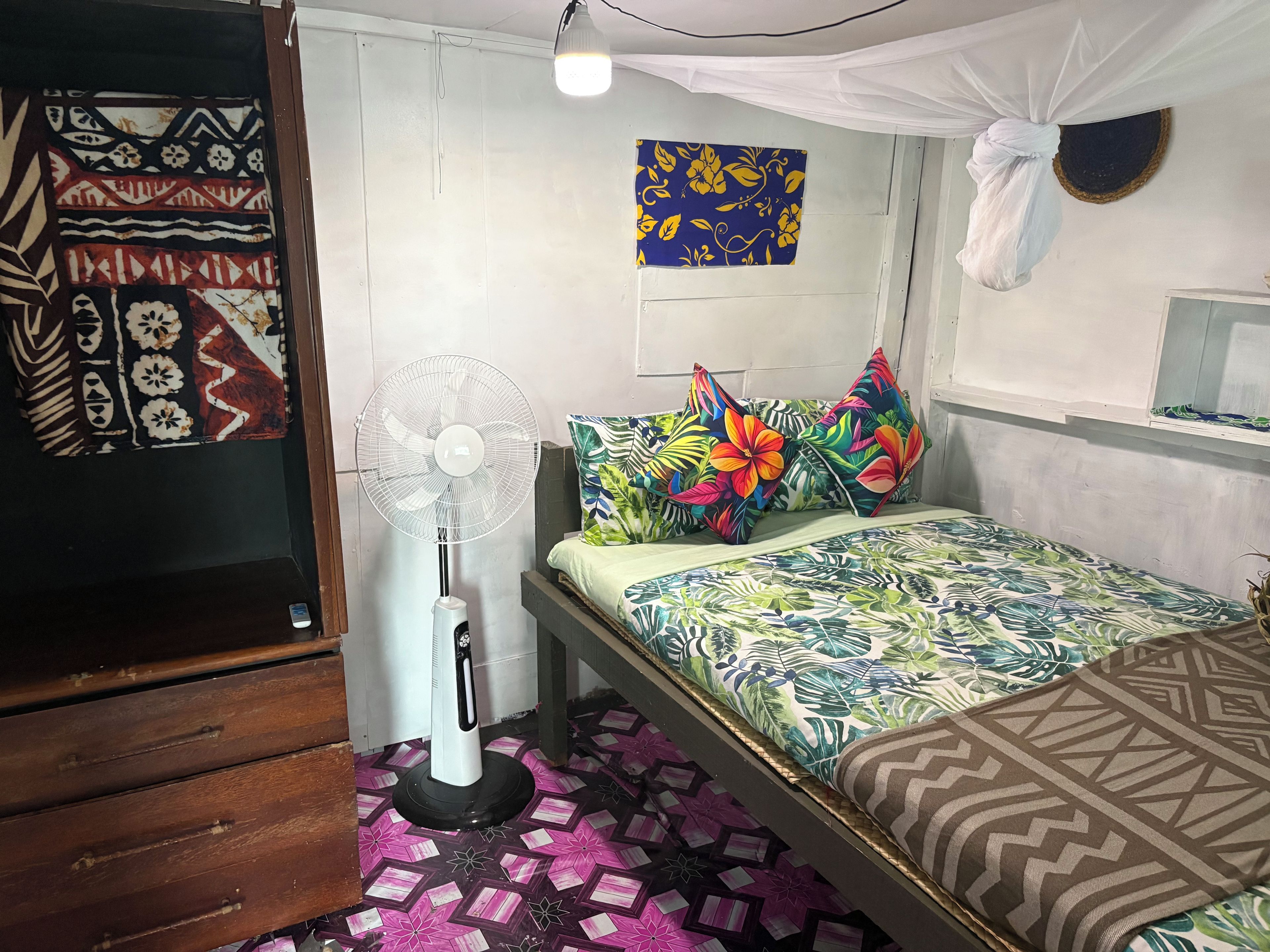 A bedroom with a bed, a standing fan, a decorative pillow, a wall hanging, and a ceiling fan.