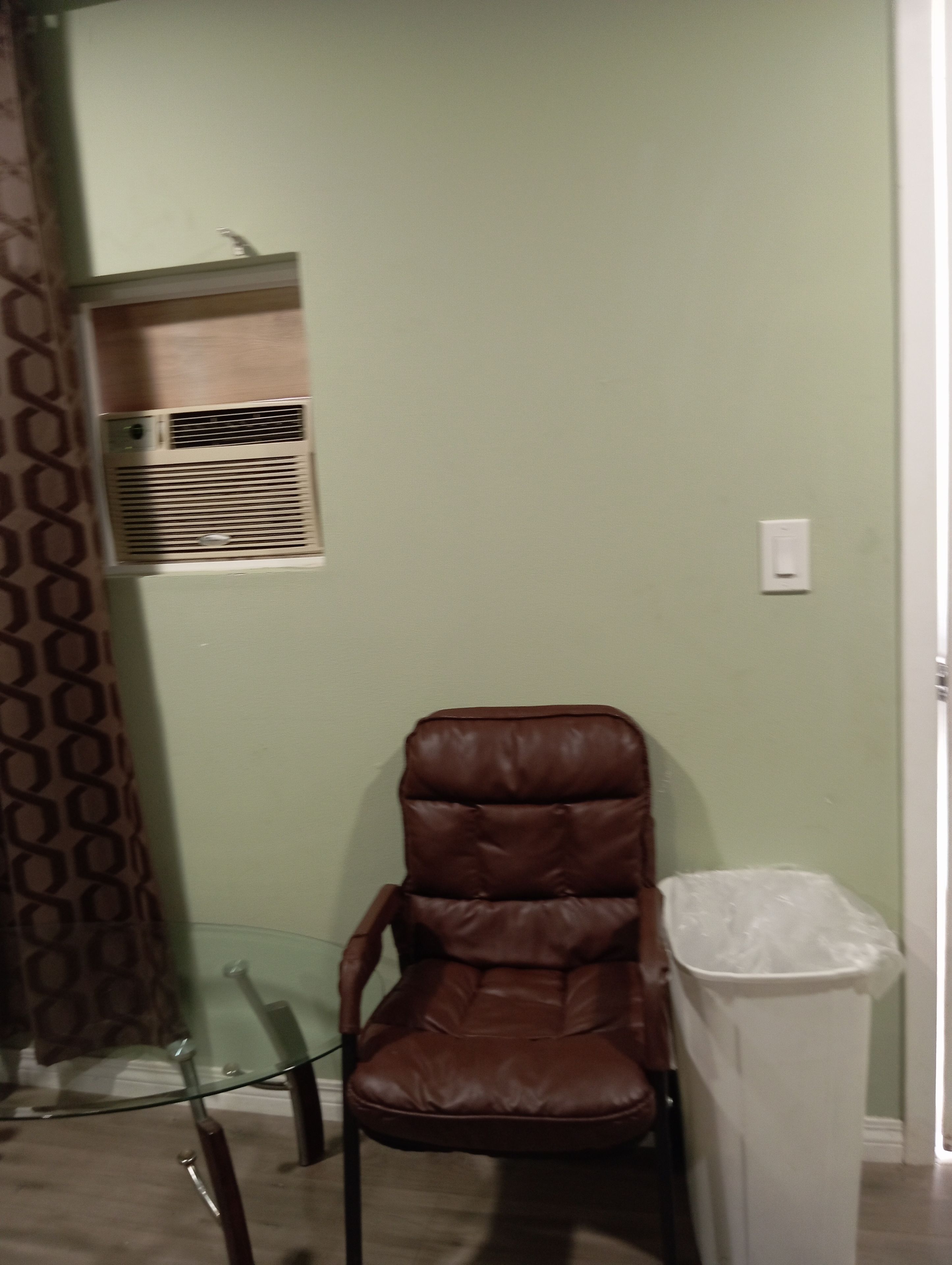 A room with a brown leather chair, a glass table, a white trash bin, and a wall-mounted air conditioning unit.