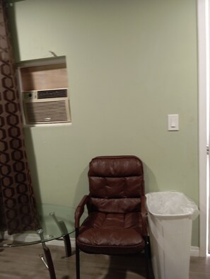 A room with a brown leather chair, a glass table, a white trash bin, and a wall-mounted air conditioning unit.