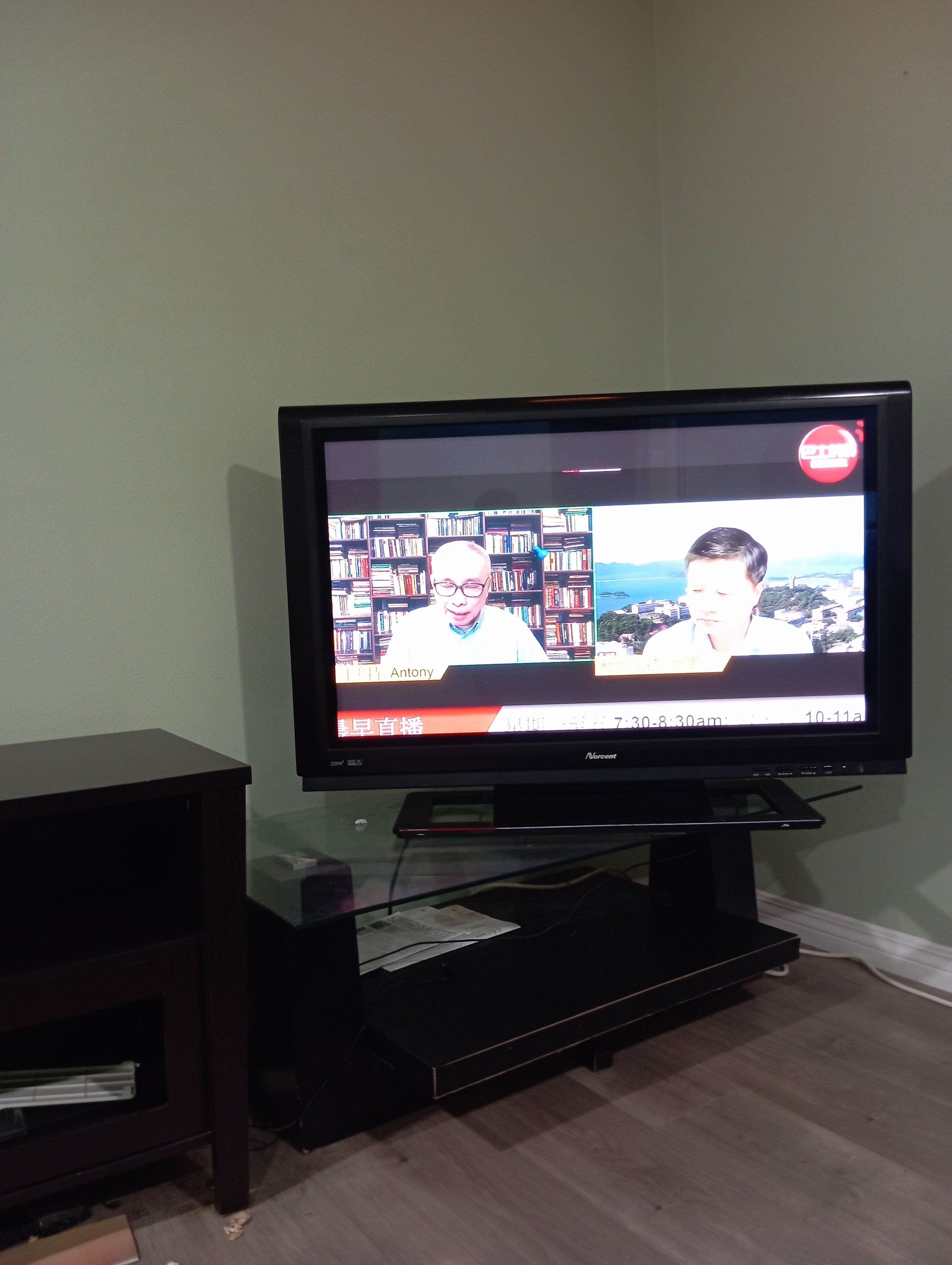 Un televisor mostrando un noticiero, con una estantería en el fondo.