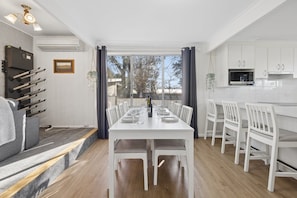 A dining area with a white table set for six, white chairs, a kitchen with white cabinets, and a view through a large window.