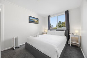 A hotel room with a large bed, a window with curtains, a bedside table with a lamp, and a radiator.