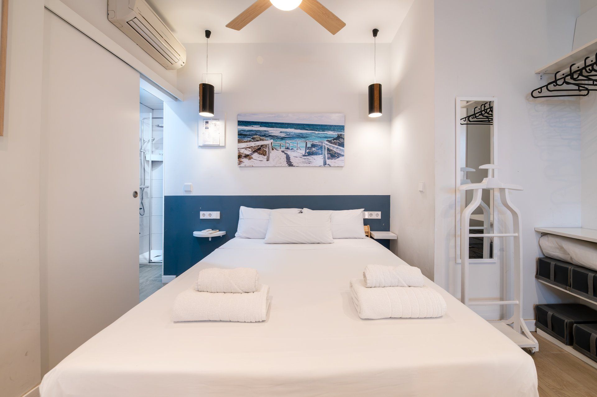 Habitación de hotel con cama, dos mesitas de noche, lámpara de noche, ventilador de techo, aire acondicionado de pared, un cuadro y un perchero.