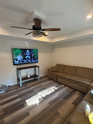 A living room with a flat-screen TV mounted on the wall, a ceiling fan, a wooden bench, a brown sofa, and a wooden floor. - The Wildcat Den @Wildcat Hideout (East Bernstadt)
