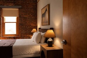 Une chambre avec un mur en briques, une grande fenêtre, un lit avec des oreillers, une lampe et une porte.