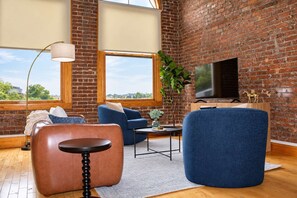 A modern living room with brick walls, a large window, a flat-screen TV, and a blue sofa. - Street View Condo | Walk to Knoxville’s Best (Knoxville)