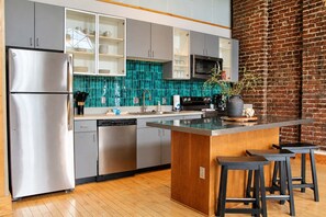Une cuisine moderne dotée d’appareils électroménagers en acier inoxydable, d’un îlot central en bois avec des tabourets, et d’un mur en briques.