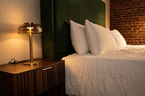A neatly made bed with a white comforter and pillows, a wooden bedside table with a lamp, and a brick wall in the background. - Street View Condo | Walk to Knoxville’s Best (Knoxville)