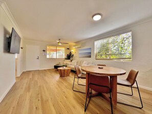 A spacious living area with a wooden floor, a round dining table with chairs, a sofa, and a TV mounted on the wall. - Swell Villa 1 the perfect coastal escape (Kalbarri)
