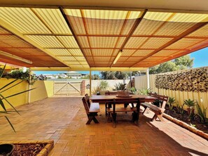 Un patio techado con una mesa y bancos de madera, rodeado de piso de ladrillo y una pared amarilla.