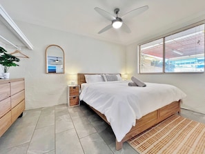 Un dormitorio con cama, ventilador de techo, mesita de noche con lámpara, espejo y ventana con persianas.