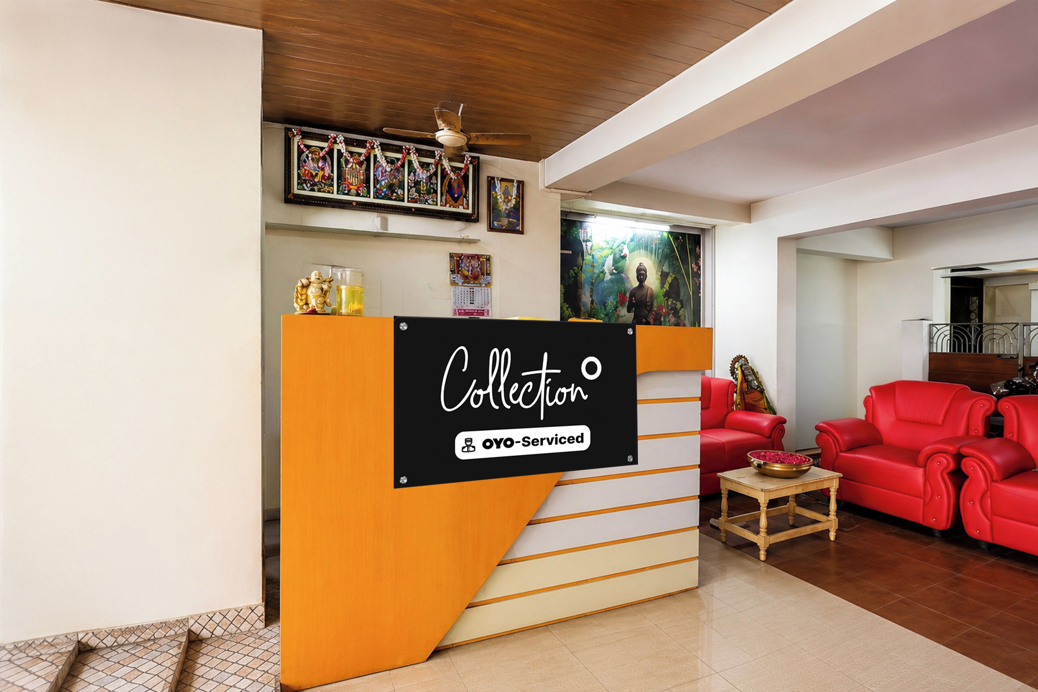 Zona de recepción con un cartel que dice 'Collector' y 'OYO-Serviced'. Hay un mostrador de recepción de madera, un sofá rojo y una pequeña mesa con un cuenco. La habitación tiene techo de madera y suelo de baldosas.