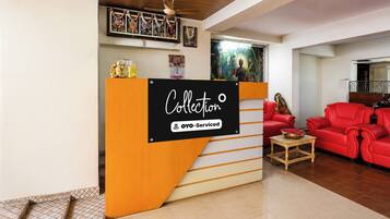 Zona de recepción con un cartel que dice 'Collector' y 'OYO-Serviced'. Hay un mostrador de recepción de madera, un sofá rojo y una pequeña mesa con un cuenco. La habitación tiene techo de madera y suelo de baldosas.