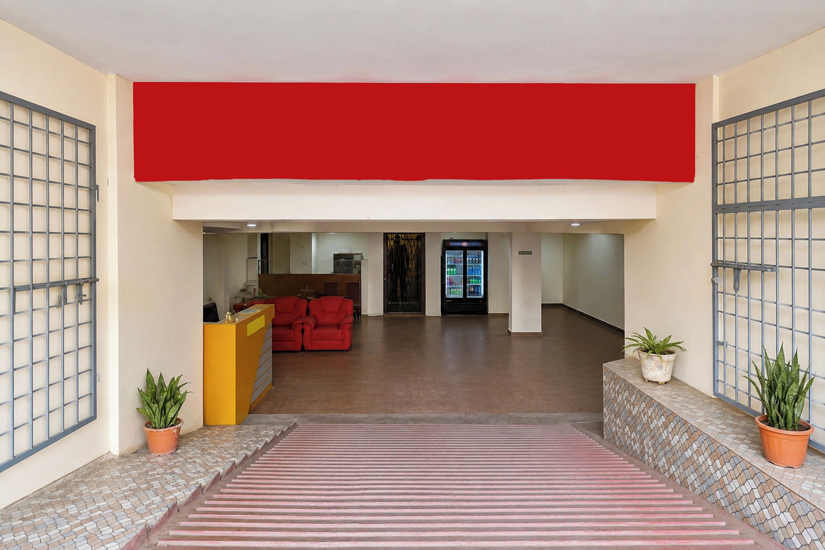 A reception area with a red wall, a reception desk, and a seating area with red chairs. There are potted plants on either side of the reception desk.