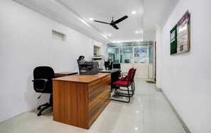 Uma área de recepção com uma mesa de madeira, cadeiras de escritório pretas, um ventilador de teto e um painel de vidro com um aviso de boas-vindas.