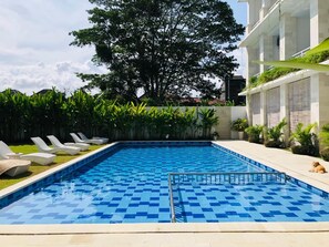 Piscina com padrão quadriculado, rodeada por espreguiçadeiras e vegetação.