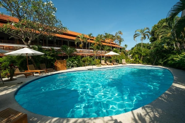 A swimming pool with lounge chairs and umbrellas, surrounded by lush greenery and a building with a tiled roof.