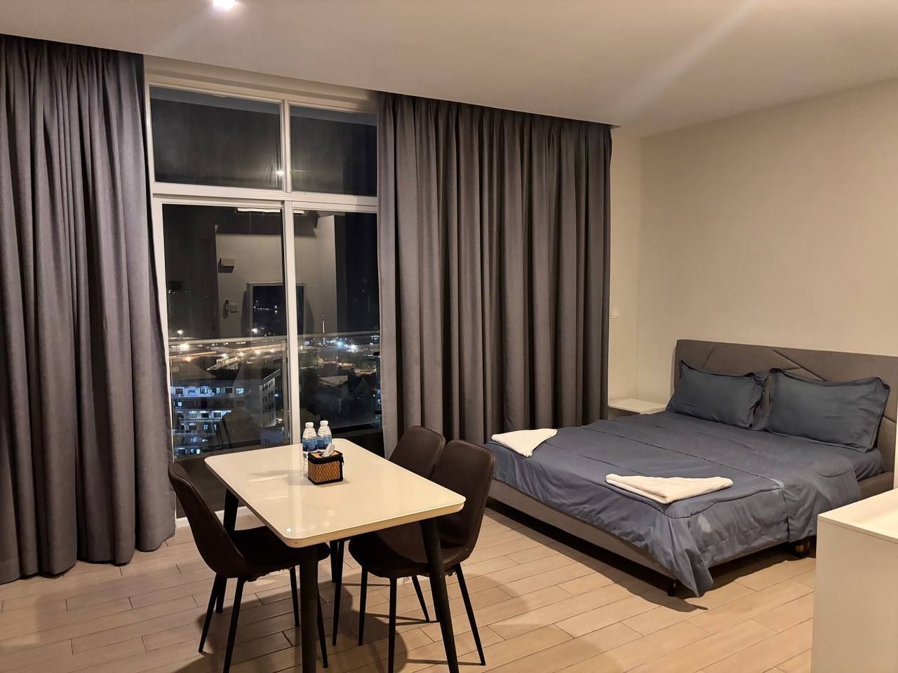 A hotel room with a bed, a dining table with chairs, and a large window with curtains.