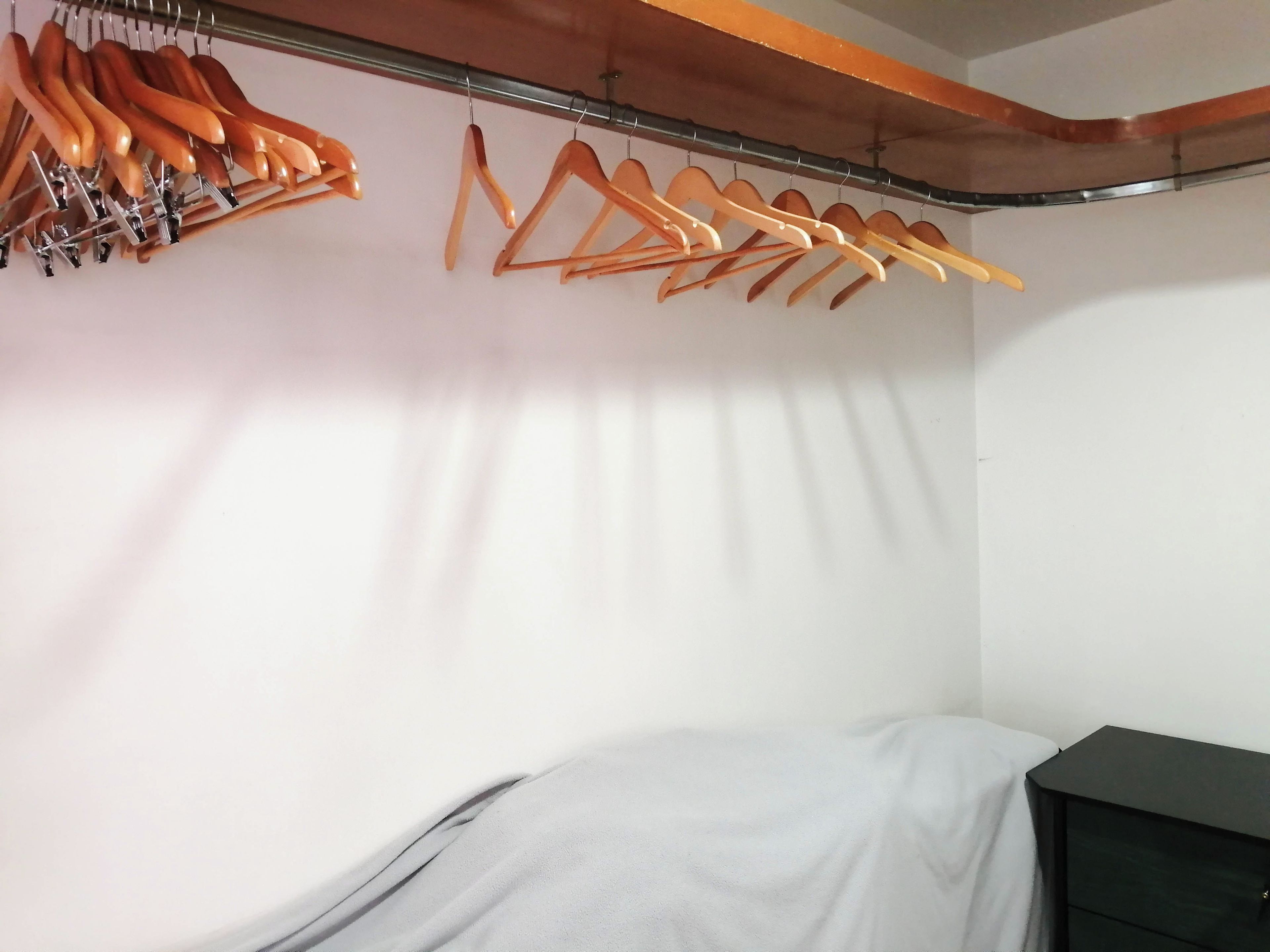 A room with a bed covered in a white sheet, a black dresser, and a clothing rack with hangers.