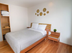 Un dormitorio ordenado con una cama de madera, una mesita de noche, un estante con almohadas y decoración en la pared.