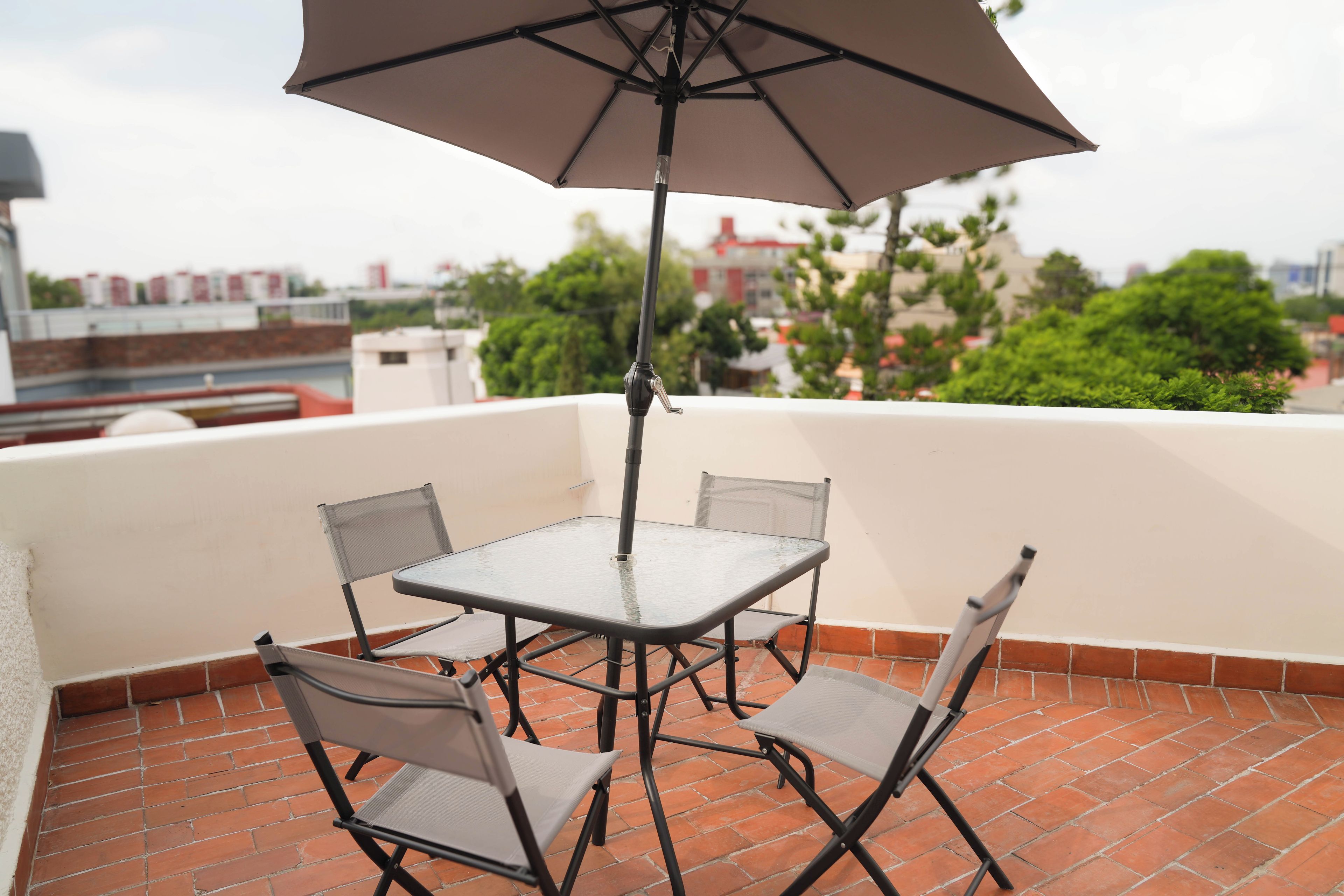 Un patio con una mesa, cuatro sillas, una sombrilla y vistas a edificios y árboles al fondo.