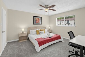 Ein Schlafzimmer mit Bett, Nachttischen, Schreibtisch, Stuhl, Deckenventilator, Fenster mit Jalousien und einem gerahmten Bild an der Wand.