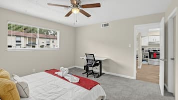 Un dormitorio con cama, escritorio, ventilador de techo, ventana con persianas y vistas al exterior.