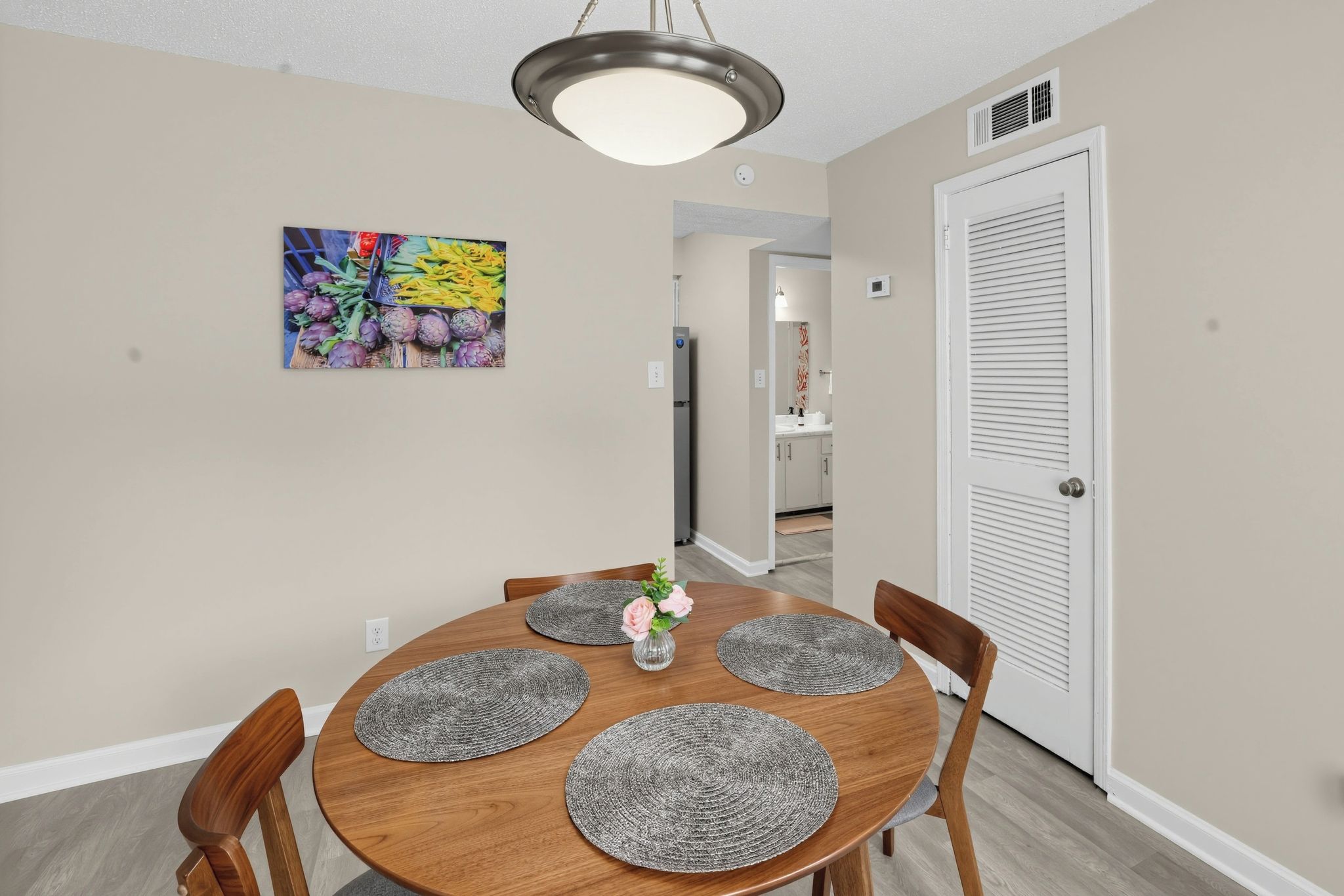 A dining area with a round wooden table, four chairs, a vase with flowers, and a ceiling light fixture.
