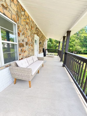 A covered porch with a stone wall, a wicker sofa, and a black railing. - Family Pet friendly Private 11-Acre Country Retreat with 6 bedrooms & Disc Golf (Griffin)