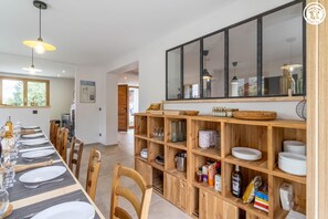 Ein Essbereich mit einem langen Tisch, der für mehrere Personen gedeckt ist, Holzregale mit Gläsern und Geschirr sowie ein großer Spiegel über den Regalen.
