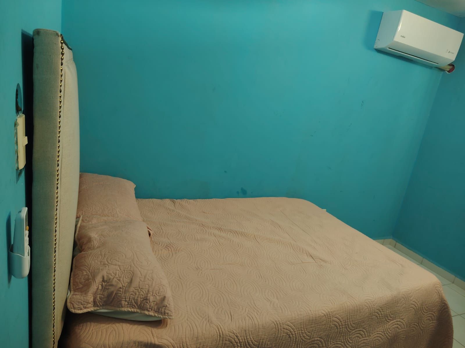 A single bed with a headboard, a wall-mounted air conditioner, and a power outlet on the wall.
