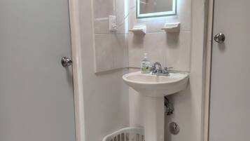 A bathroom with a pedestal sink, a mirror, a hand dryer, and a trash bin.